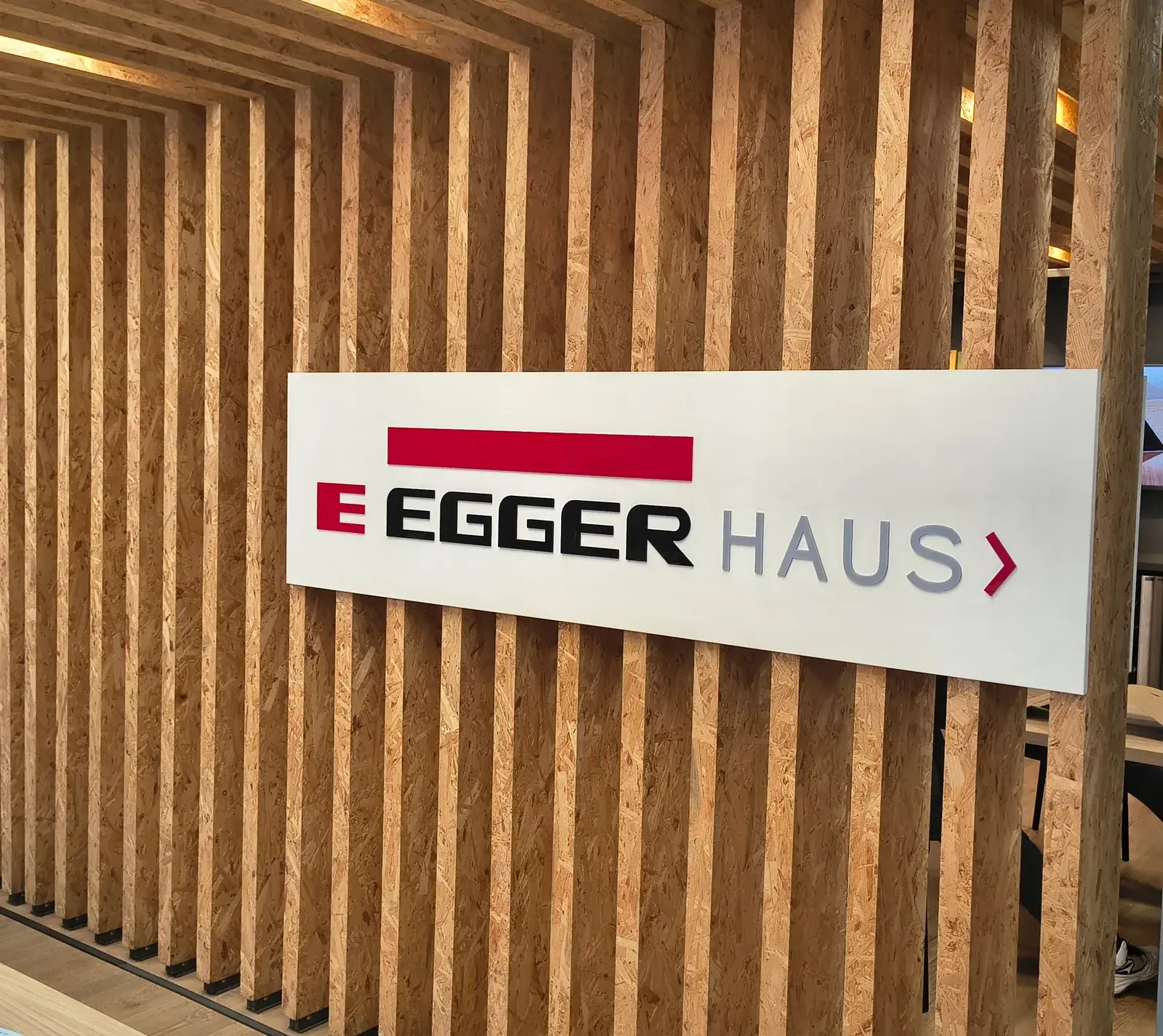 Cartel de chapa blanca con el logo EGGER HAUS montado sobre una pared de listones verticales de madera.