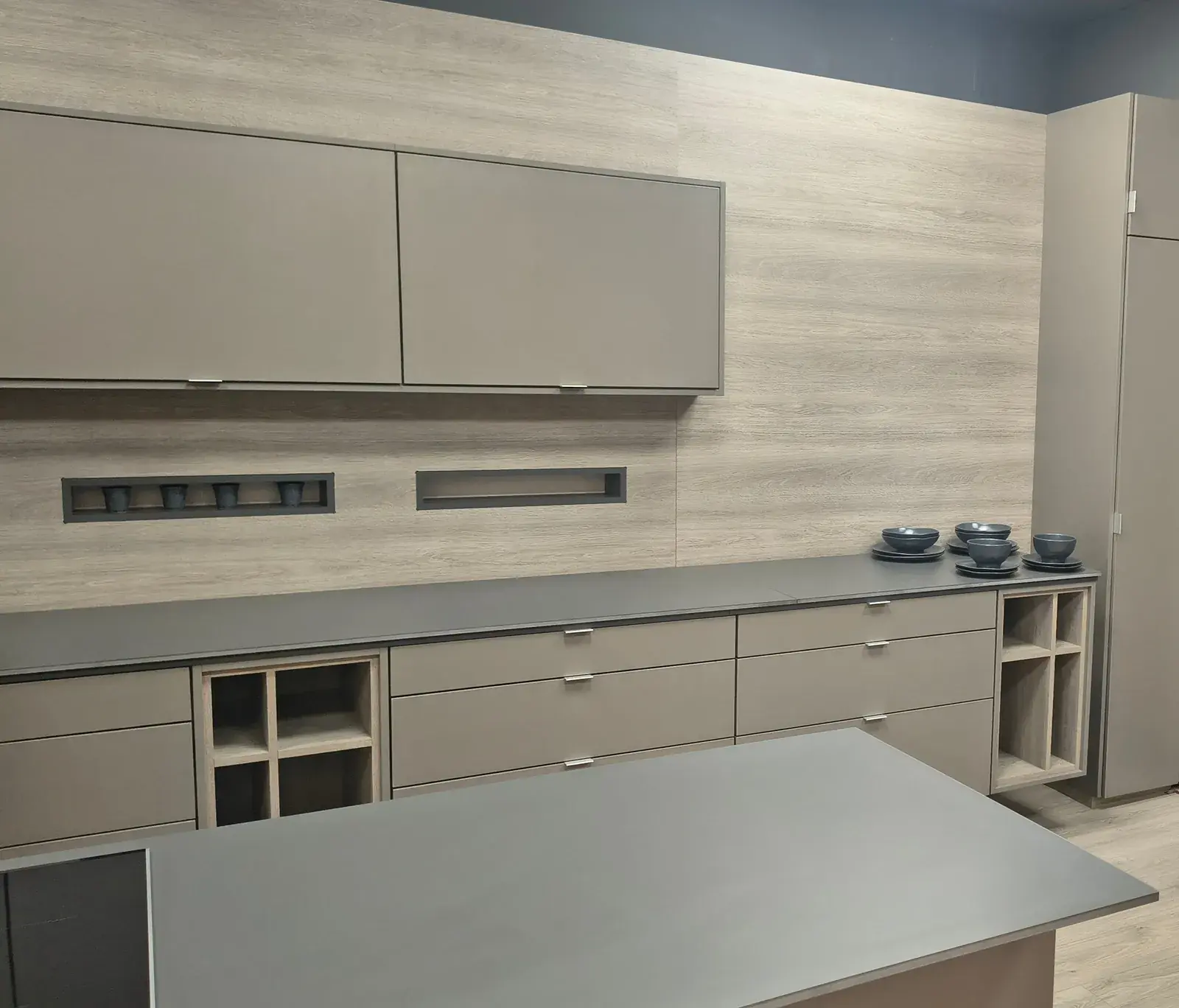 Cocina completa en el showroom: alacenas y bajomesada en gris topo, isla central y revestimiento de melamina símil madera clara.