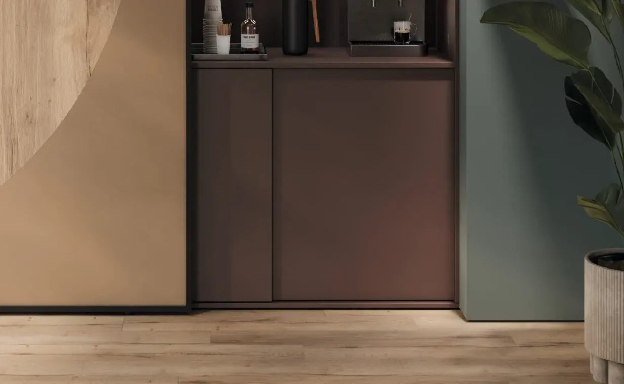 Mueble bajo de puertas corredizas color chocolate con una cafetera y botellas encima, entre un panel de madera clara y uno verde, con una planta al costado.