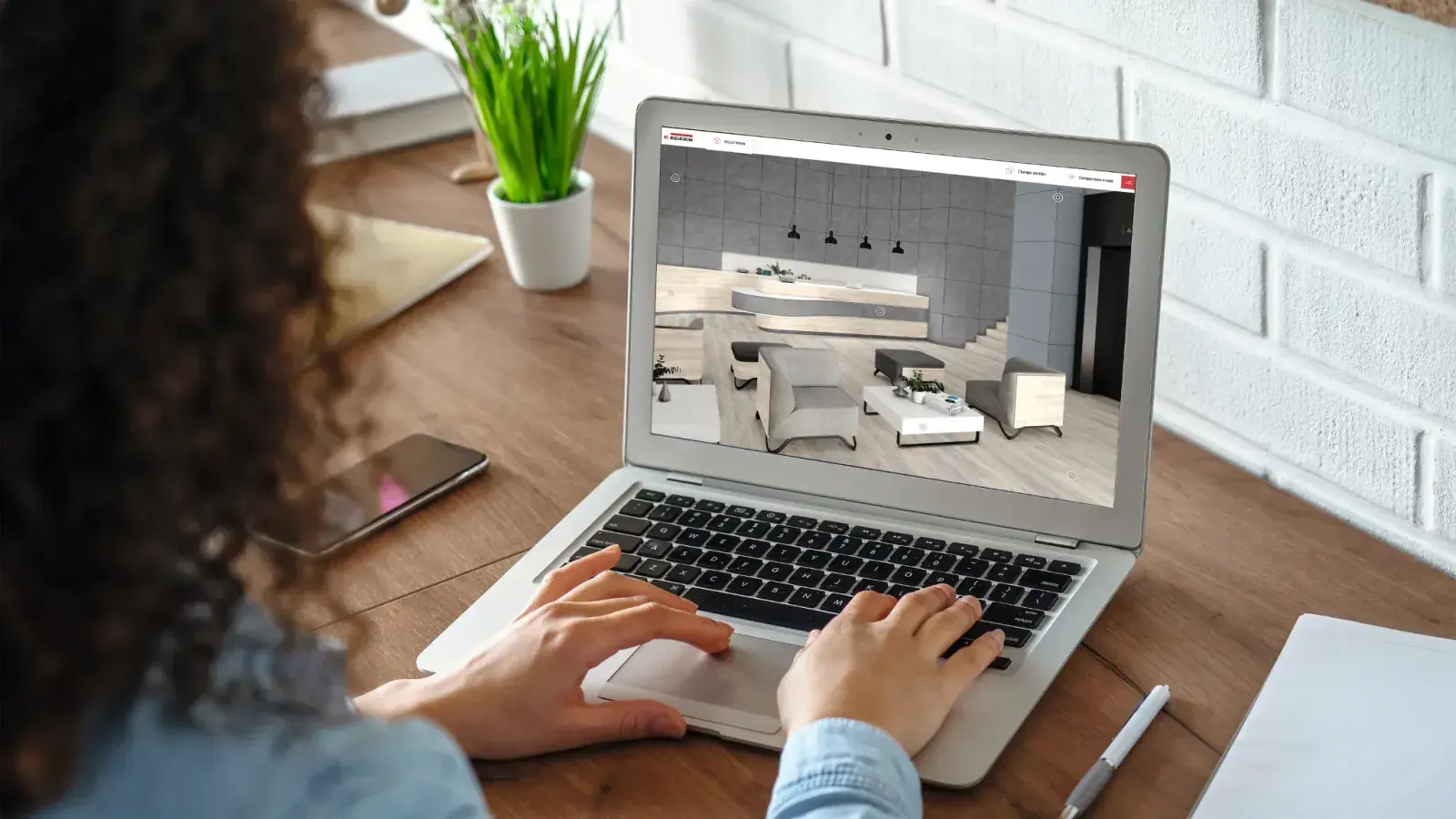 Una persona trabaja en una notebook con el Estudio de Diseño Virtual de EGGER abierto: en la pantalla, un ambiente en 3D con piso de madera clara, una barra curva y sillones grises.