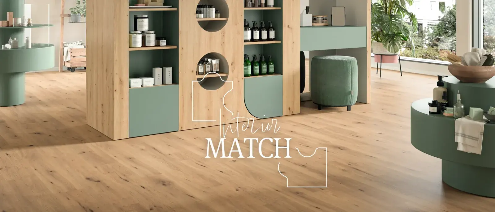 Ambiente de tienda resuelto con Interior Match: un mueble expositor de madera clara con frentes verde salvia, un tocador y un puf del mismo verde, y en todo el piso una madera clara que repite la veta del mueble. Sobre la foto, el logotipo Interior Match.