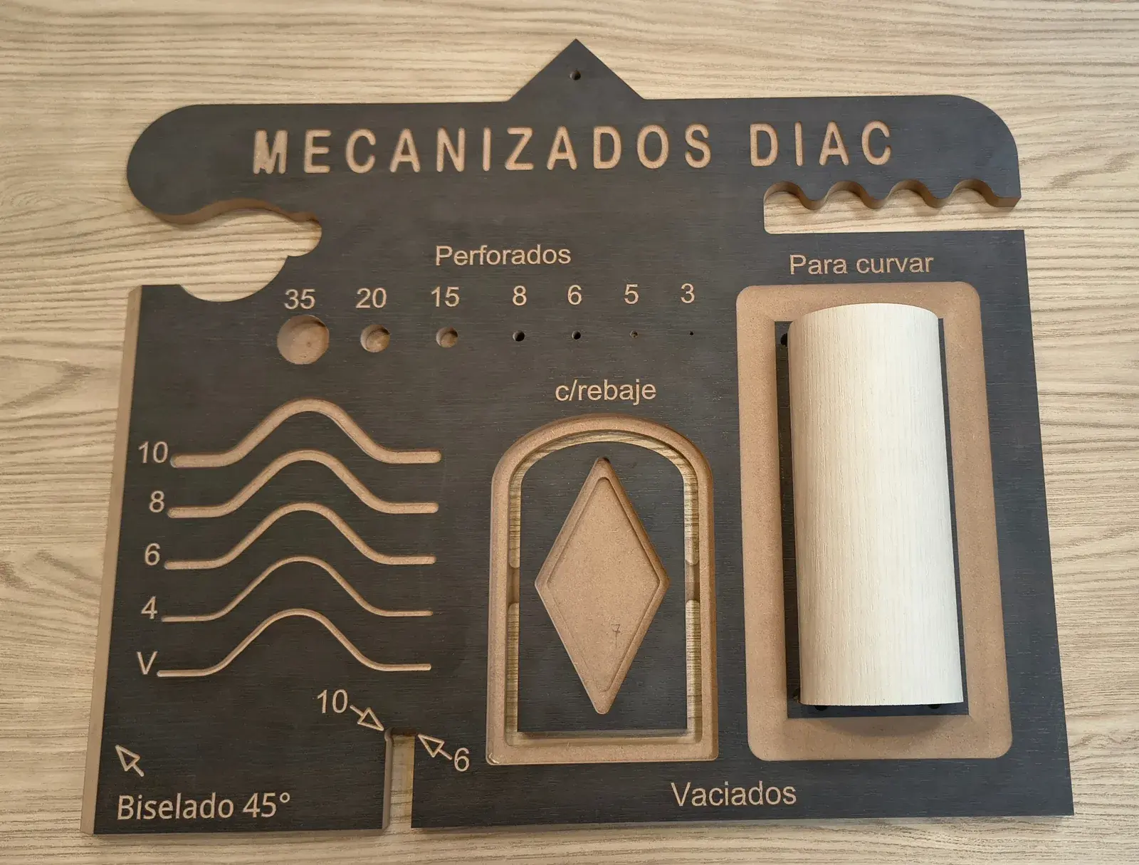 Muestrario de mecanizados hecho por DIAC: una placa con el rótulo fresado "MECANIZADOS DIAC" que reúne un ejemplo de cada trabajo — perforados de 3 a 35 mm, ranuras de 4 a 10 mm y en V para plegar, biselado a 45°, vaciados con rebaje, cantos ondulados y una pieza curvada.