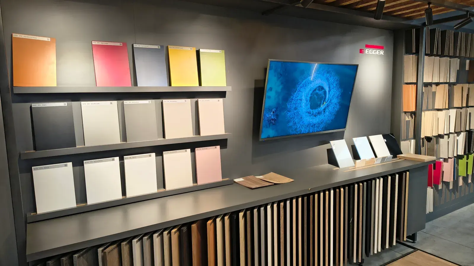Pared del EGGER Studio con las muestras de placas unicolor en tres estantes —naranjas, rojos y azules arriba, grises y blancos abajo—, cada una rotulada con su código, y a la derecha los muestrarios de maderas colgados junto a una pantalla con el logo de EGGER.